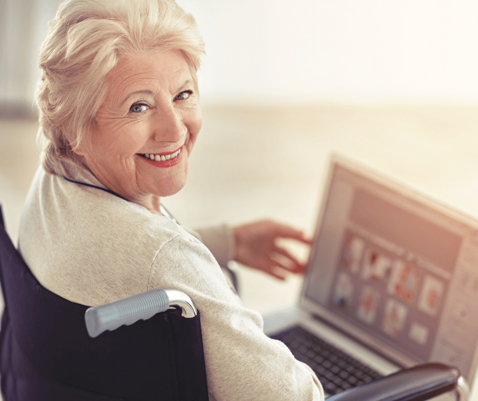 An older white woman in a wheelchair points to a laptop in her lap while smiling at the audience. She represents the concept of 