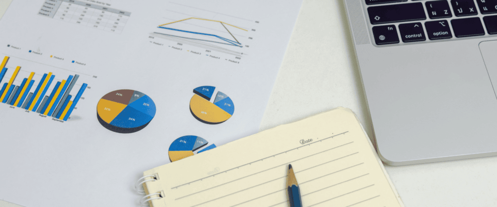 An office desk with a laptop; spreadsheet with pie, bar, and line graphs, and an empty notebook with a pencil laying atop. Represents the concept of a B2B marketing agency.