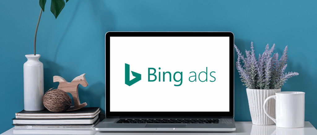 A laptop showing the Bing Ads/Microsoft Advertising/Microsoft Ads logo setting on a desk, set against a blue background.