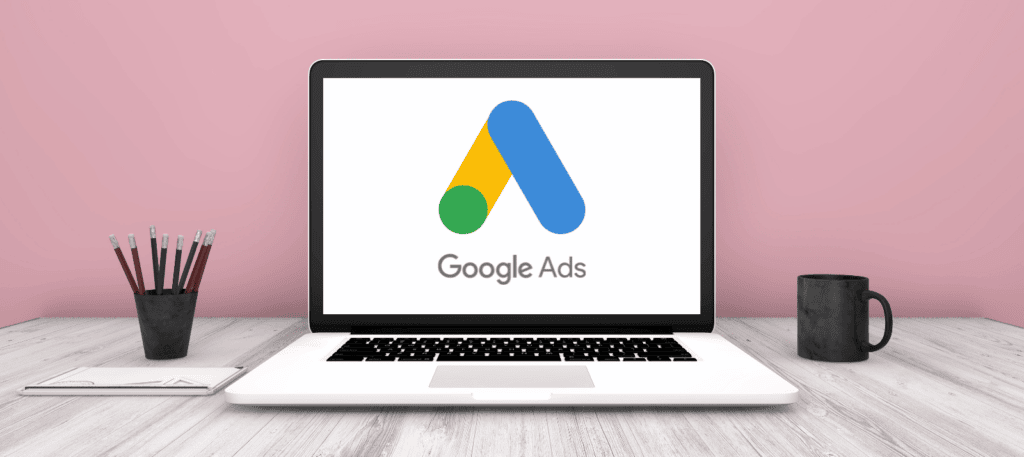 A laptop with Google Ads onscreen sits on a desk, set against a pink background.