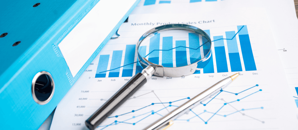 A magnifying glass and pen lay overtop a pile of spreadsheets tracking product sales.