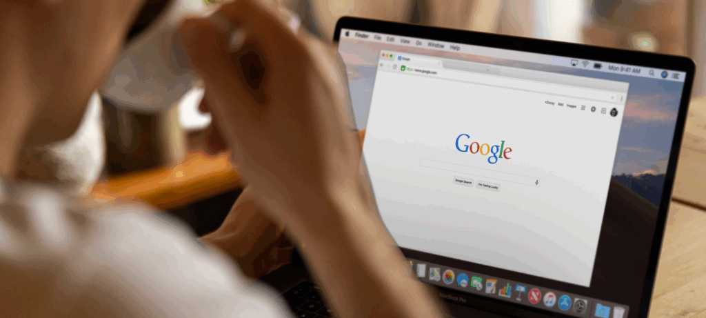 Person drinking coffee works on a laptop with Google Search pulled up.