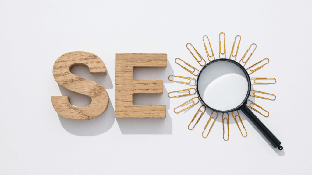 Against a white background, two block letters "S" and "E" are next to a magnifying glass surrounded by paperclips. It spells "SEO"