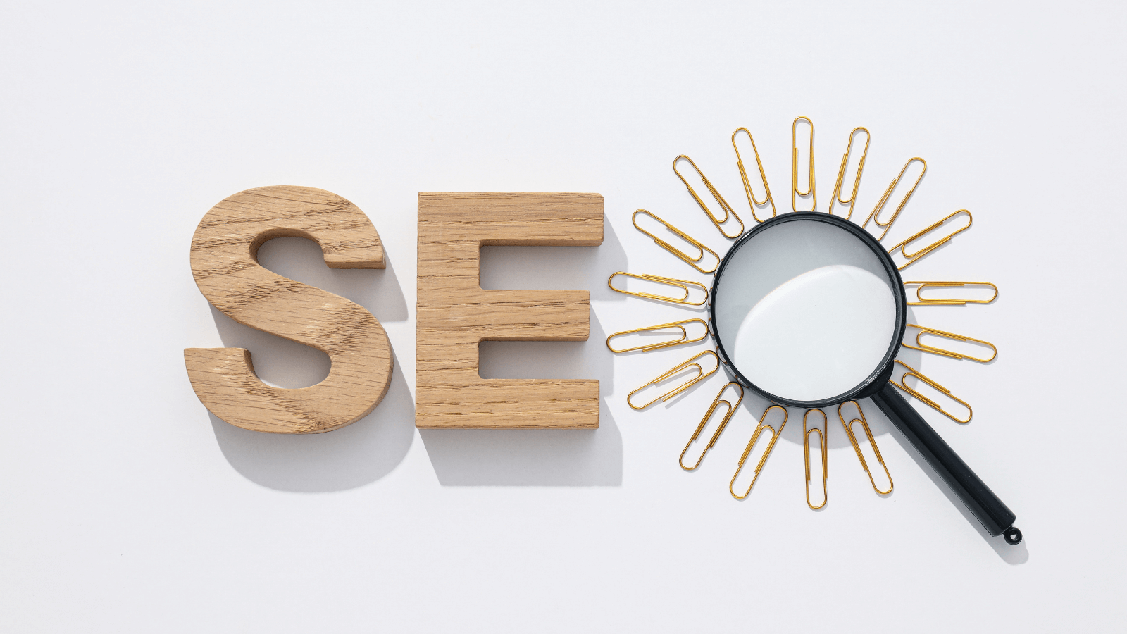 Against a white background, two block letters "S" and "E" are next to a magnifying glass surrounded by paperclips. It spells "SEO"
