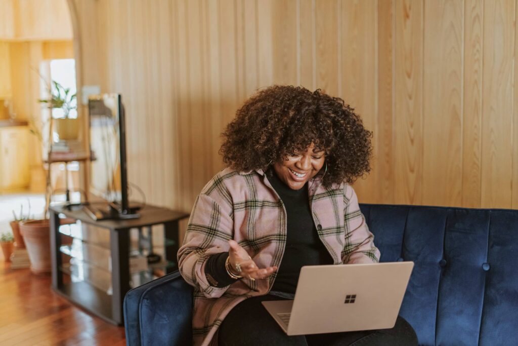 A woman on a couch laughs while using a laptop. Photo by Surface on Unsplash.